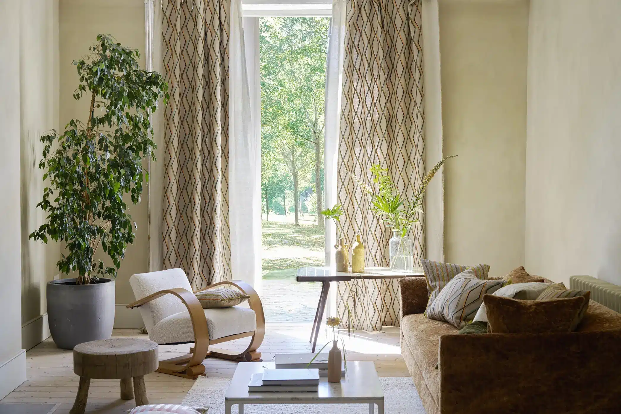 Wohnzimmer mit Raumausstattung: braunes Sofa, weißer Sessel, Zimmerpflanze und lange Fenstervorhänge.