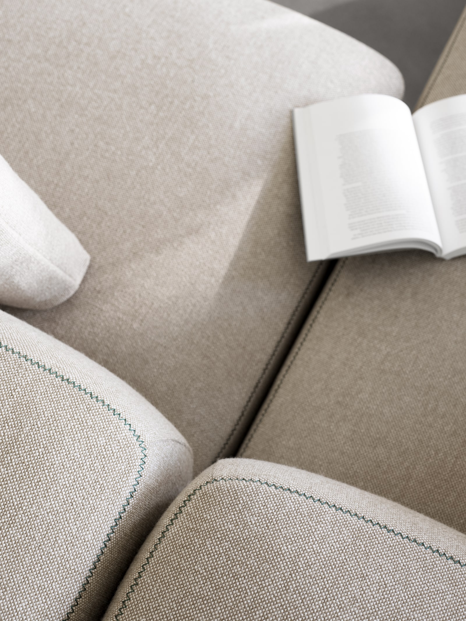 Nahaufnahme von beigen Polsterkissen mit grüner Naht, offenem Buch und Fenster-Sichtschutz im Hintergrund.